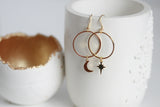 Gold hoop earrings with star and moon charms on a white textured surface.