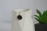 A gold circular pendant with a black center charm, hanging from a small gold chain, displayed on a white faceted ceramic vase next to a green succulent.
